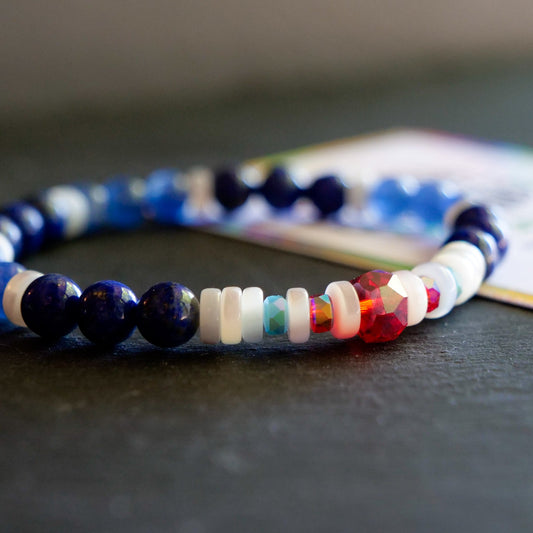 A beaded stretch bracelet featuring dark blue lapis lazuli, white beads, light blue beads, and a red faceted crystal charm, resting on a dark surface with a blurred card in the background.