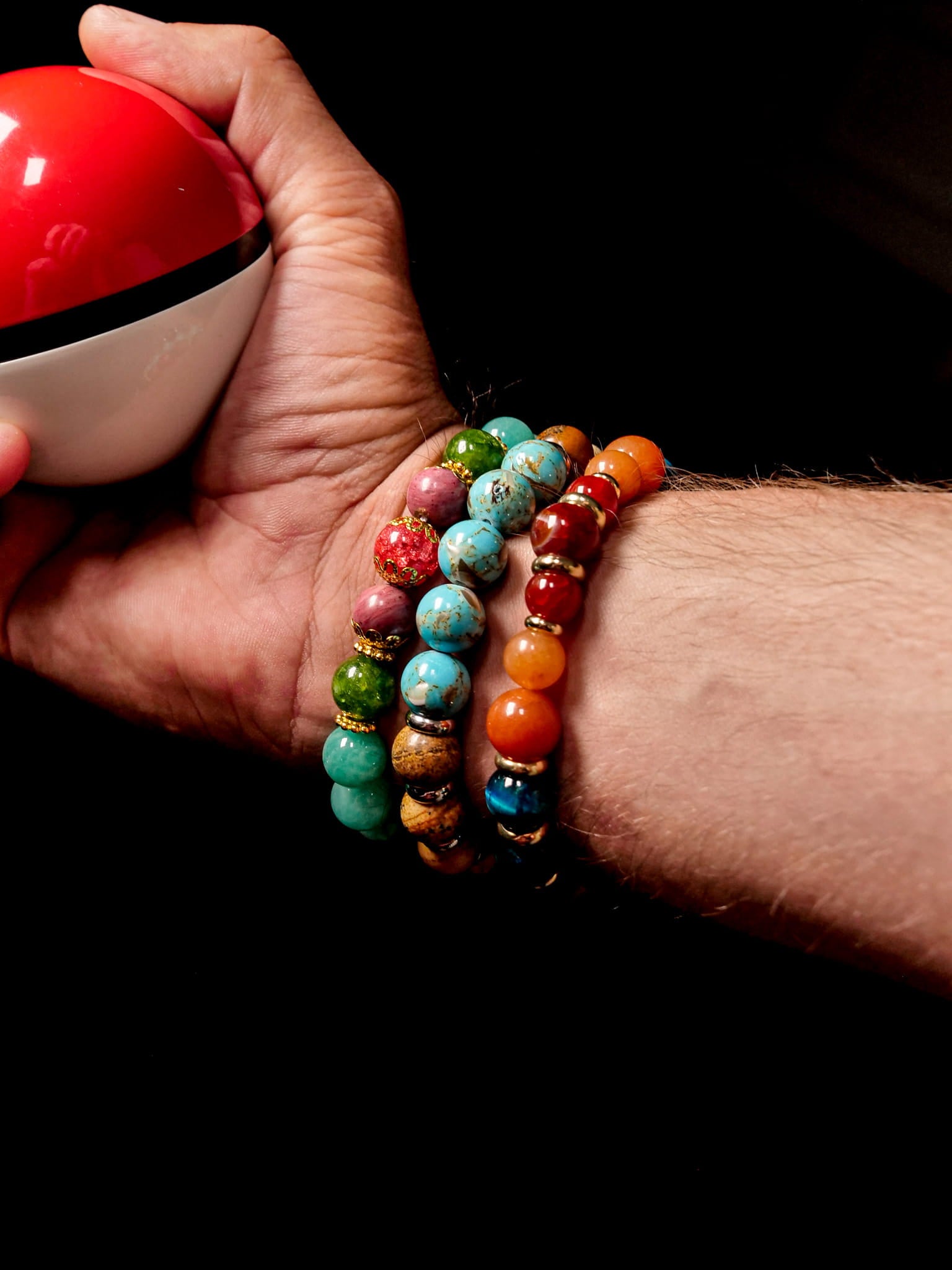 A close-up of a person's wrist wearing three colorful beaded bracelets, with one hand holding a red and white Pokéball against a black background.